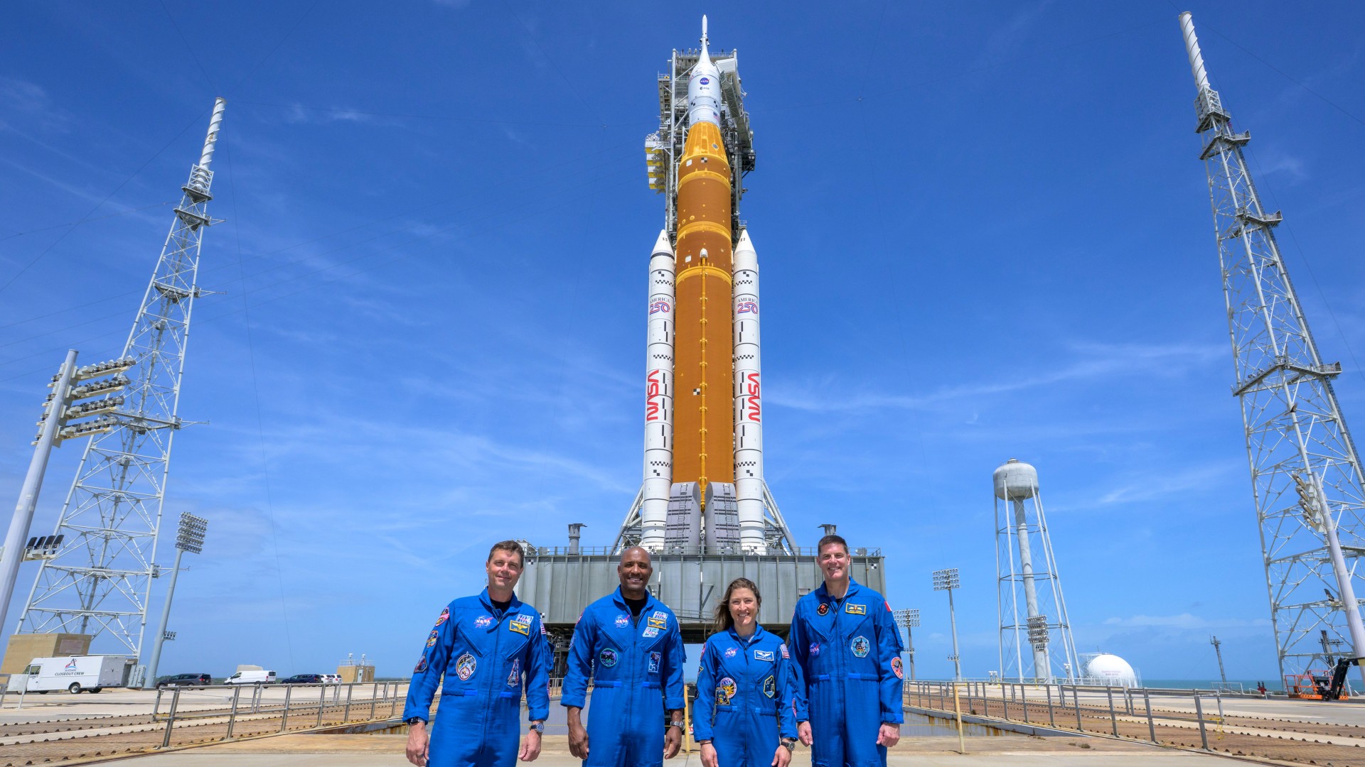 four astronauts in blue jumpsuits posing with a giant nasa rocket