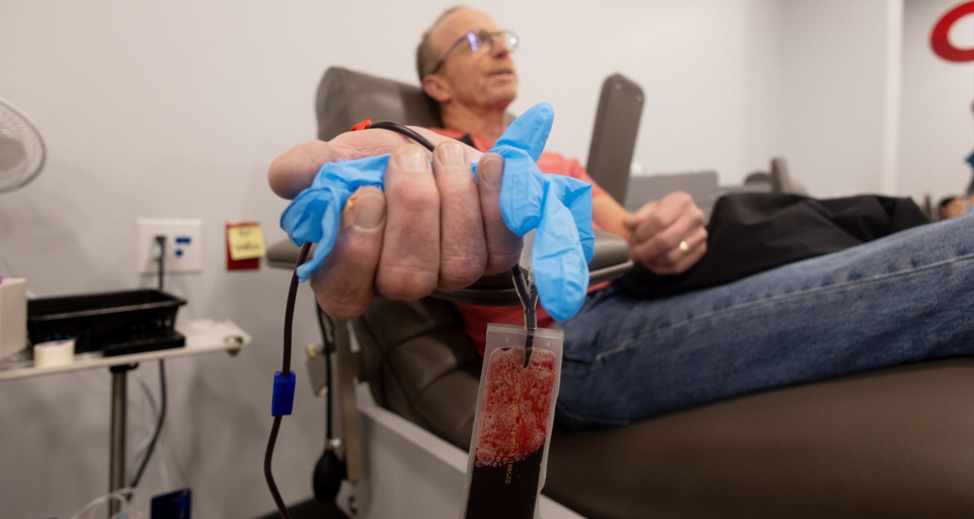 A person reclines in a medical donation chair during a blood or plasma donation, with one hand in the foreground holding a blue glove around tubing connected to a clear collection bag partially filled with dark red blood. Medical equipment and a clinical room setting are visible in the background.