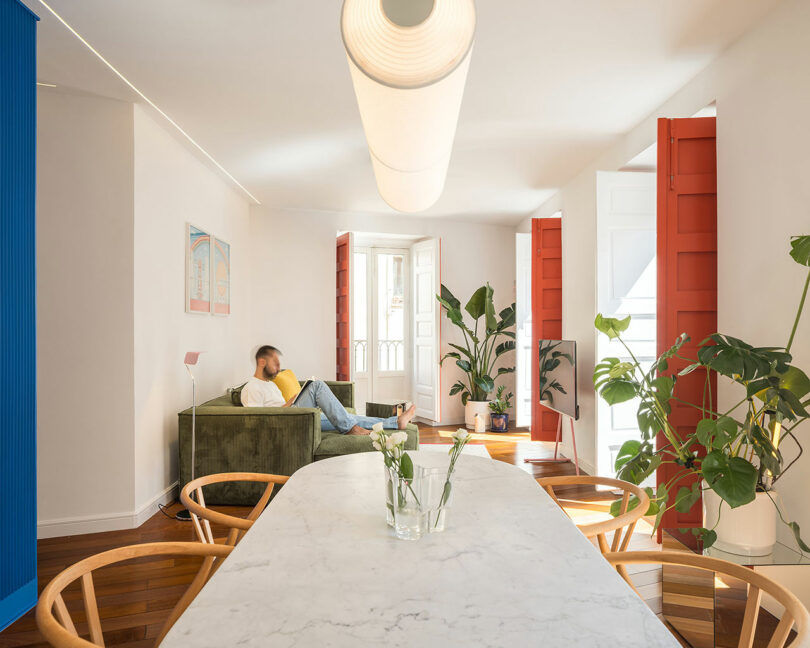 A man sits on a green sofa in a bright living room with red window shutters, houseplants, wooden chairs, and a marble dining table with a glass vase.