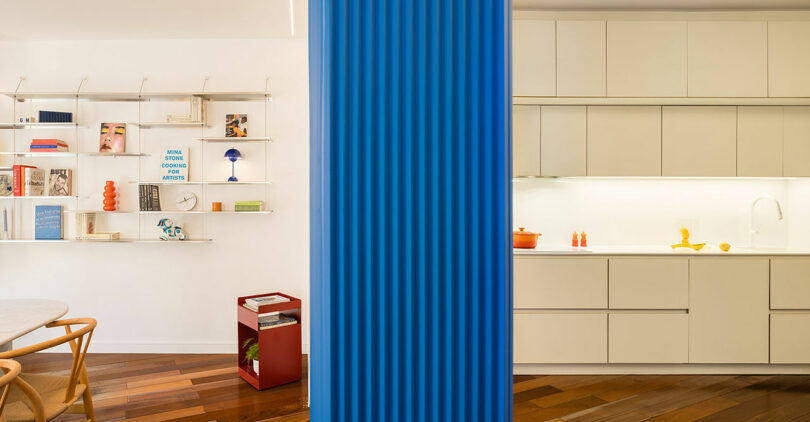 Modern interior with beige kitchen on the right, open shelving with books and decor on the left, separated by a vertical blue ribbed divider.