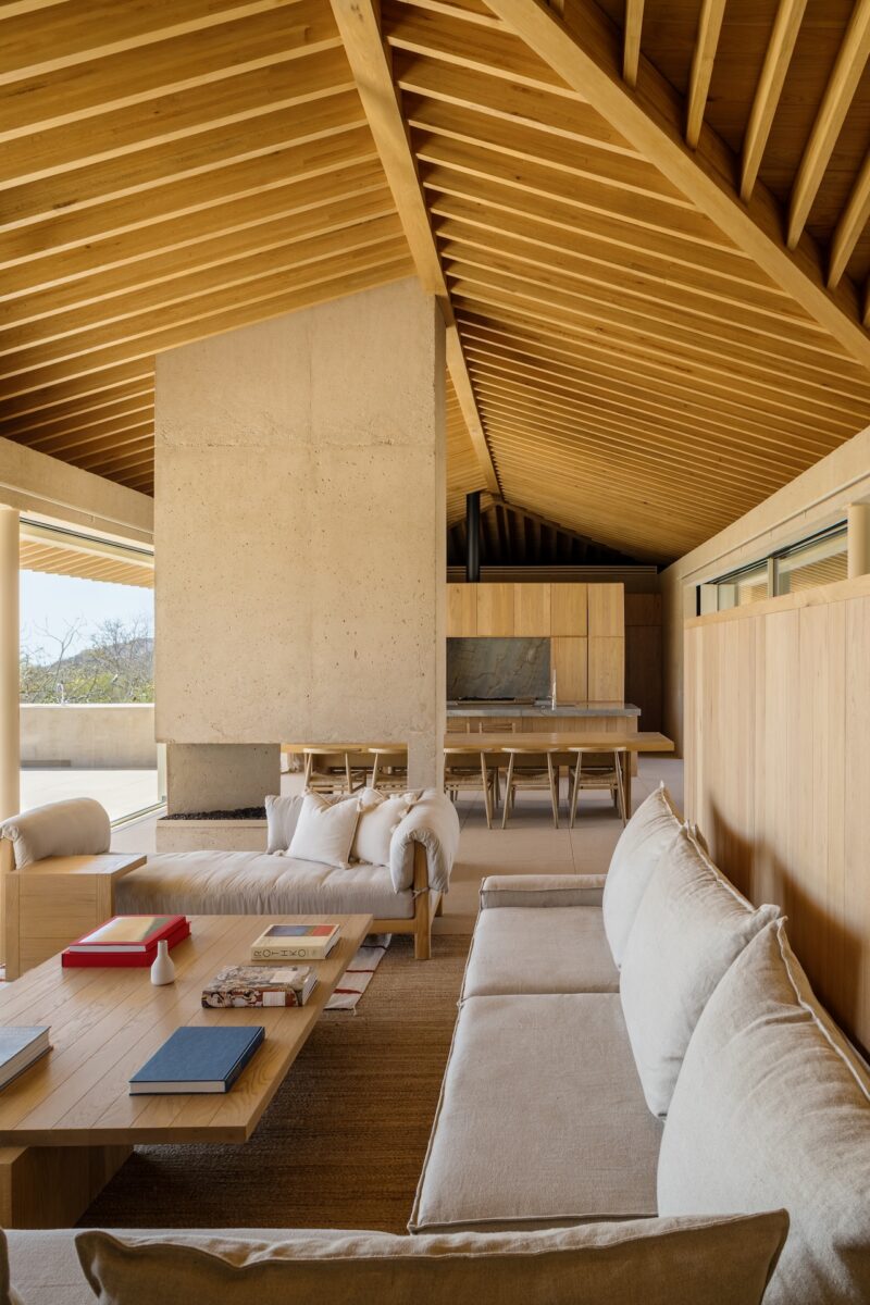Modern living and dining area with light wood beams, neutral-toned sofas, a large concrete fireplace, and a wooden dining table, all featuring minimalist design elements.