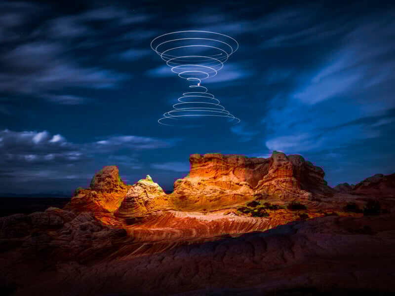 Red rock formations are illuminated under a night sky, with swirling white light trails hovering above, creating a mesmerizing spiral pattern. Some clouds drift across the sky, and the unique landscape stands out dramatically.