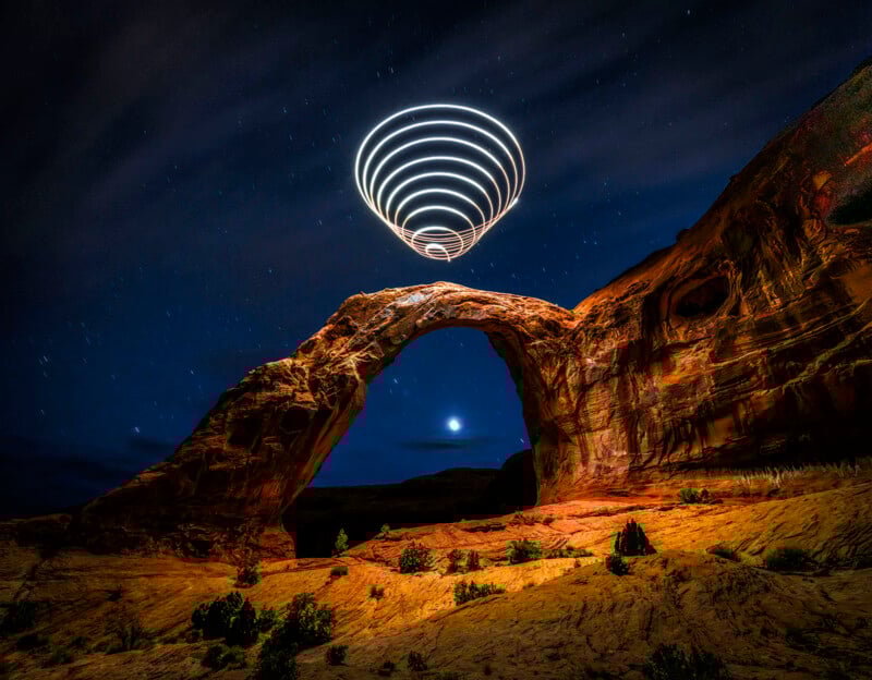 A natural rock arch under a starry night sky, with glowing spiral light trails above it, creating a futuristic effect. The landscape is illuminated, highlighting red rock formations and scattered vegetation.