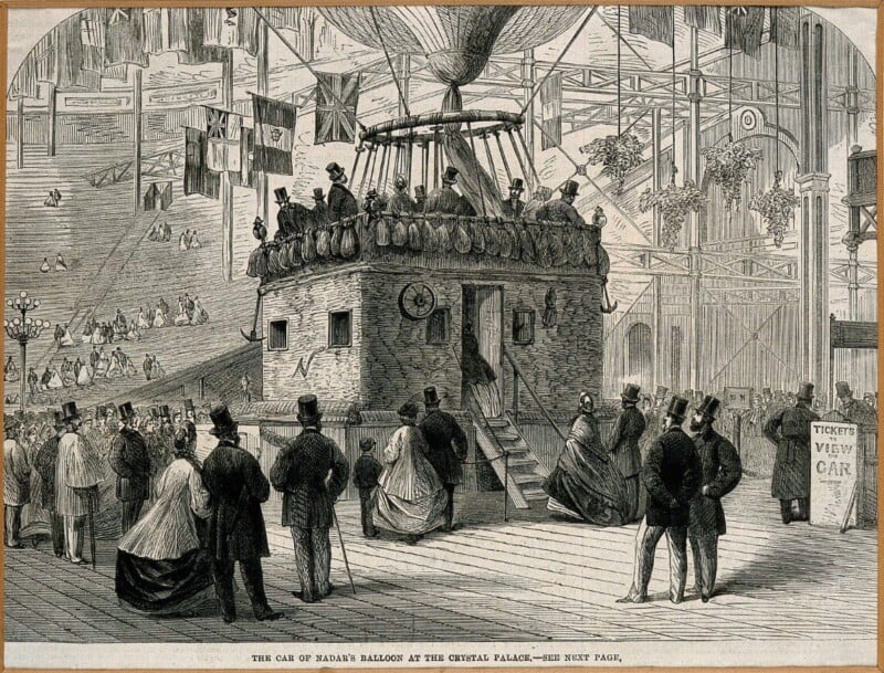 An old illustration shows people gathered around a large balloon’s car on display in an ornate indoor exhibition hall. Onlookers in 19th-century clothing stand nearby; some are onlookers, others board the car. Flags and banners hang overhead.