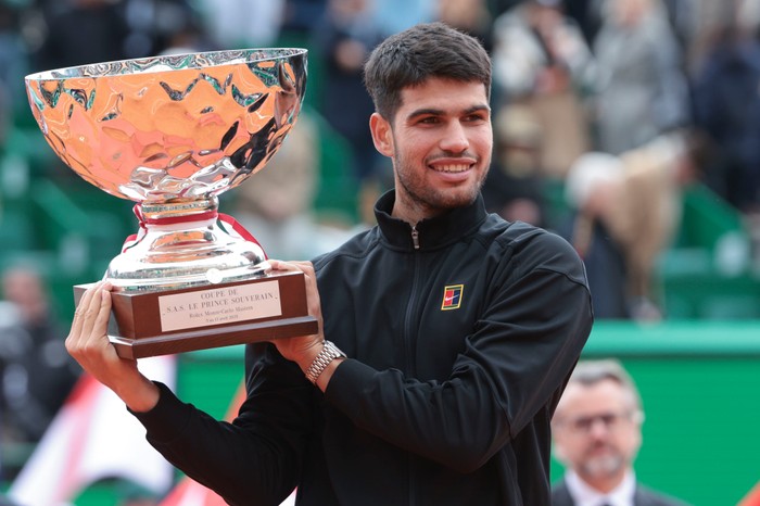 Tennis player Carlos Alcaraz, in a black Nike jacket, smiles as he holds up the Monte Carlo Masters trophy on court.
