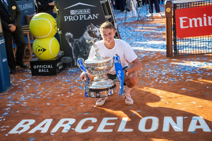 Holger Rune Danish tennis player Holger Rune, in a white t-shirt, crouches and smiles on the court at the Barcelona Open with the trophy in his hands.