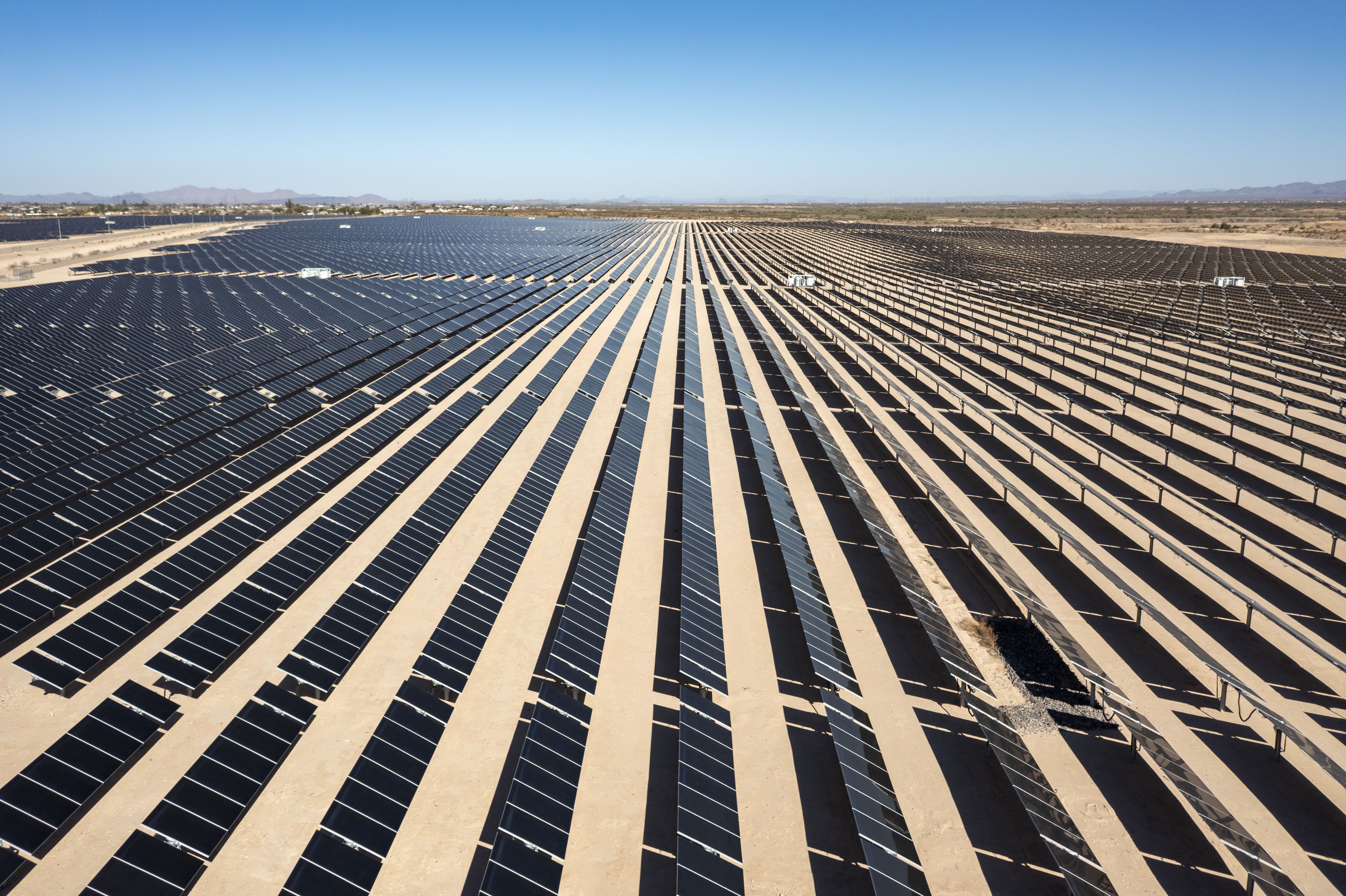 A vast solar panel farm stretches across an arid desert landscape with mountains visible on the horizon under a clear blue sky.