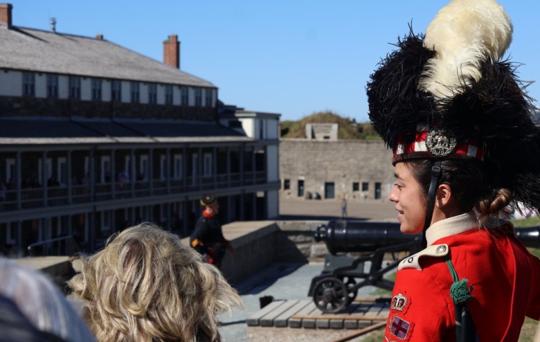 A young smiling woman in regimental uniform with a red surge and a hat with black and white feathers and a red, white, and black tartan band talks to tourists while other young guides in military uniforms get ready to fire a cannon.