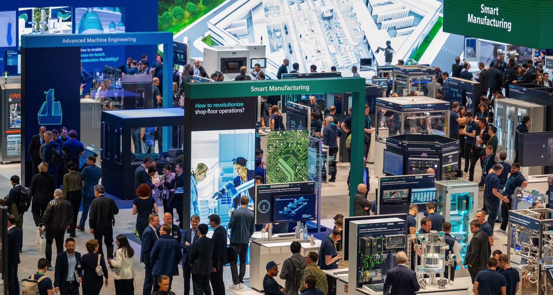 A high-angle shot of one of the pavilions at Hannover Messe, showing people lingering at a variety of robotics and technological display booths. Signs reading "Advanced Machine Engineering" and "Smart Manufacturing" hang from the ceiling.