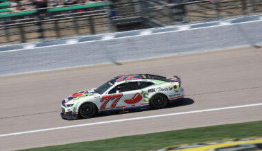 White race car number 77 speeding along a track with blurred spectators in the stands behind it