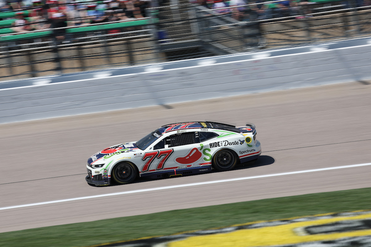 White race car number 77 speeding along a track with blurred spectators in the stands behind it