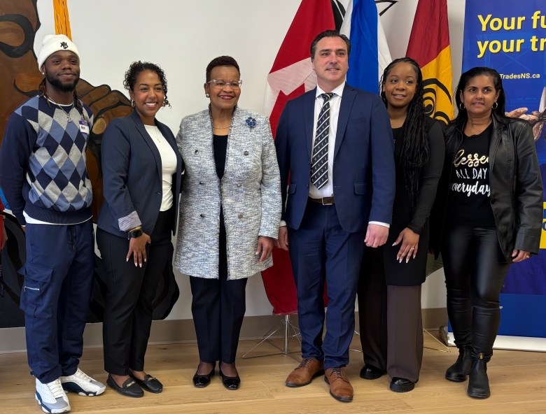 A man with a white hat, blue and grey checkered shirt with navy pants, stands next to a woman in a navy blazer and a white shirt. Next to the woman is an older woman with a tweed grey coat and clear glasses. Then there is a man in a navy suit with brown hair, and next to him is a woman with cornrows, a black long sweater and brown pants. Beside her is a woman with a leather jacket, leather pants, and a shirt that says "blessed all day, everyday." They are all smiling for a picture.