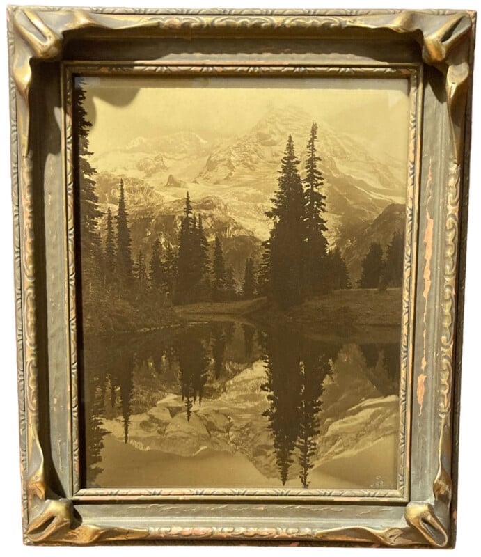 Sepia-toned framed photograph of a mountain landscape, featuring tall pine trees and a still lake reflecting the trees and peaks. The vintage frame has ornate, worn gold detailing.