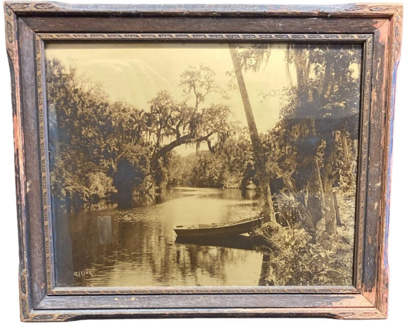 Sepia-toned photo in a rustic wooden frame shows a calm river surrounded by trees with moss. A small rowboat is tied to a leaning tree on the riverbank in the foreground.
