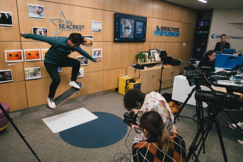 A skateboarder performs a trick indoors as two people photograph him, with cameras and recording equipment set up in a media studio decorated with photos and Seacrest Studios signs.