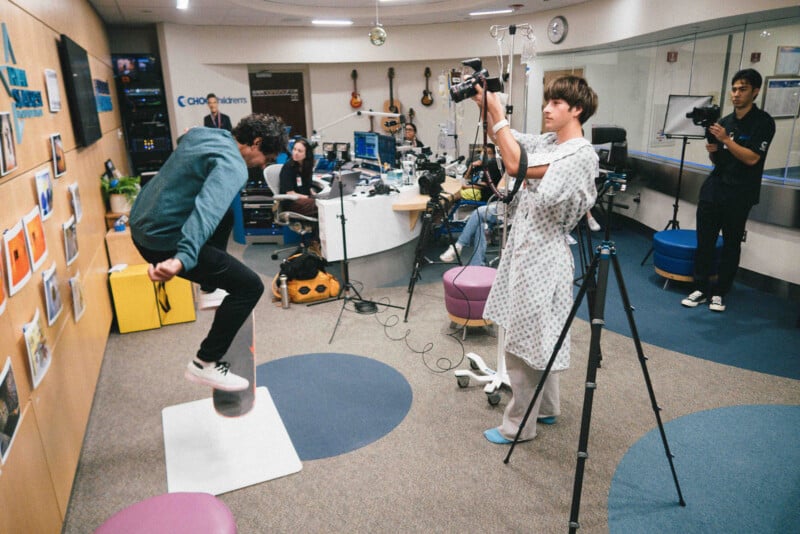 A person in a hospital gown films another person jumping on a balance board in a colorful hospital room, with cameras and equipment set up and staff visible in the background.