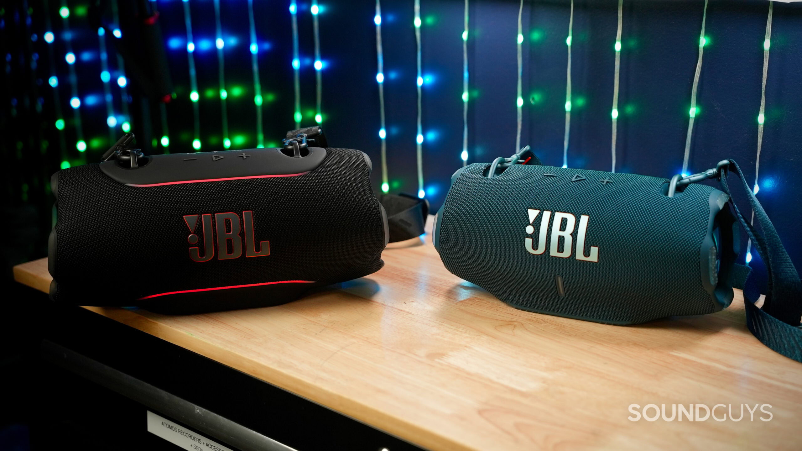 JBL Xtreme 5 vs Xtreme 4 - 1 (1) The JBL Xtreme 5 and JBL Xtreme 4 Bluetooth speakers on a wooden desk with LED lights in the background.