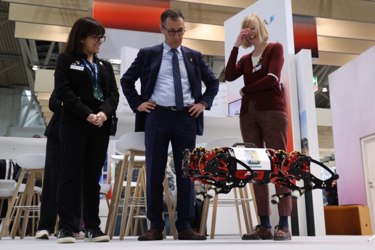 Two women and a man in a trade-show pavilion watch a robot the size of a large dog jumping about a foot in the air. The man has his hands on his hips and one of the women is laughing.