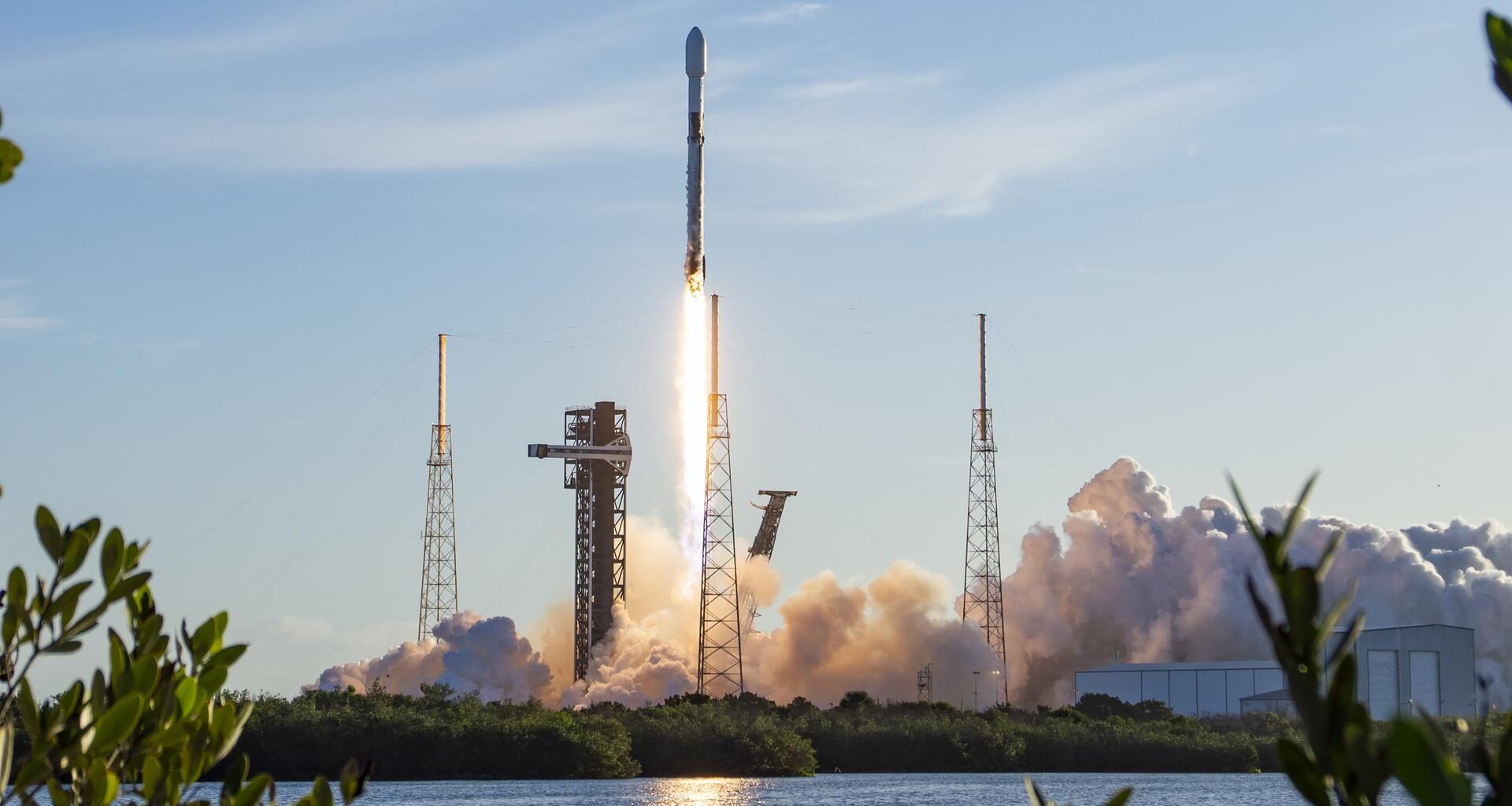Image shows a rocket launching vertically in a morning blue sky with white clouds at Cape Canaveral Space Force Station in Florida on Saturday, April 11, 2026. The launch reflects in a nearby body of water and shrubs. Photo credit: SpaceX