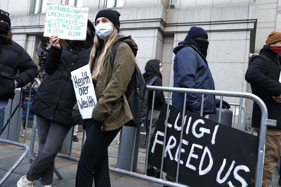 Photo of protesters in front of a barricade with signs reading Health over Wealth and Luigi Freed Us.