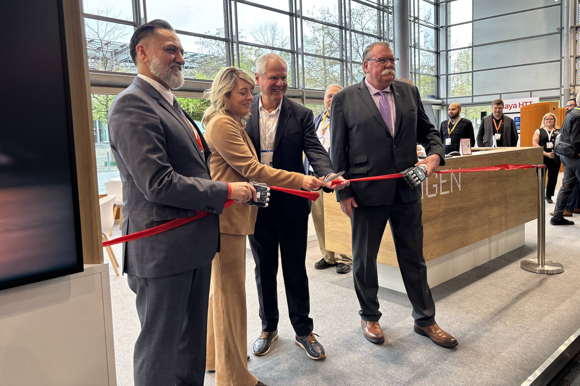 A shot of Mélanie Joly cutting a ribbon with three other people in front of a large bank of windows. Two men in the photo are holding what appear to be detached robot hands the red ribbon running through them.
