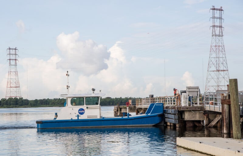 A small blue and white NASA boat is docked at a pier on calm water, with a person standing on the dock. Two tall power line towers are visible in the background against a partly cloudy sky.