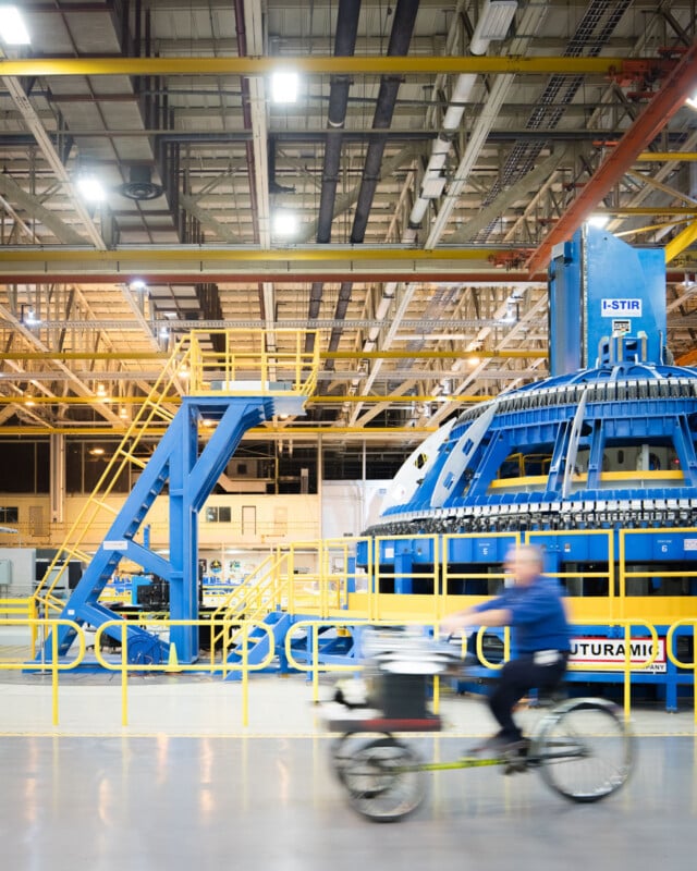 A person rides a bicycle indoors past large blue industrial machinery and yellow railings in a spacious, brightly lit factory.