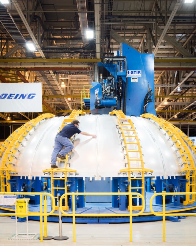 A worker stands on a yellow ladder inspecting or working on a large, dome-shaped metal structure inside an industrial facility, with machinery labeled "Boeing" and "I-STIR" in the background.