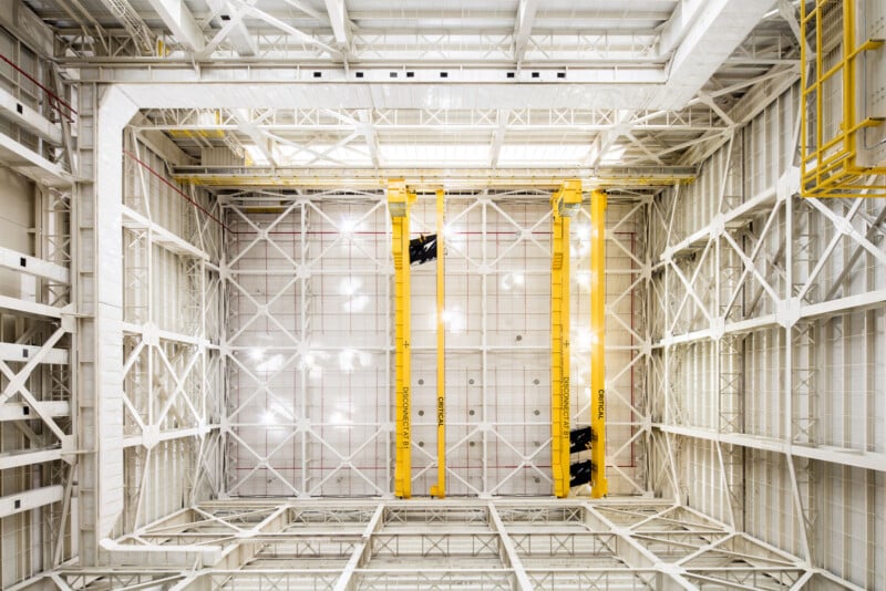 Interior view looking up at a large industrial building with white metal beams, yellow overhead cranes, and a high, grid-like ceiling structure. Bright lighting illuminates the spacious, geometric environment.
