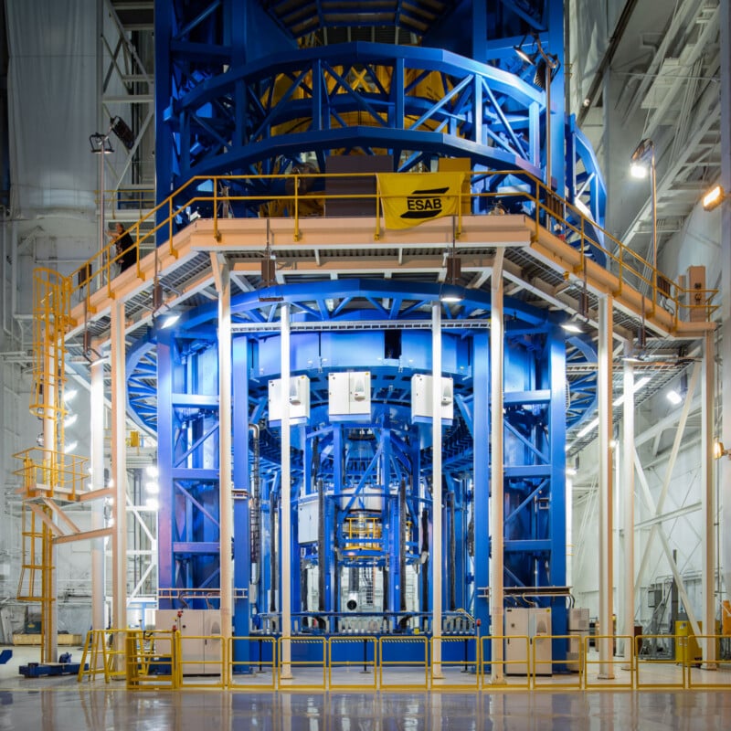 A large, blue metal industrial structure stands inside a brightly lit facility surrounded by yellow railings, platforms, and ladders, with various pipes and equipment visible.