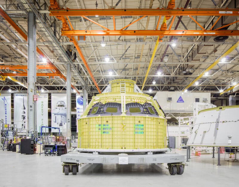 A spacecraft module with a yellow exterior is displayed on a platform inside a spacious, brightly lit industrial facility with high ceilings, metal beams, machinery, and NASA logos visible in the background.
