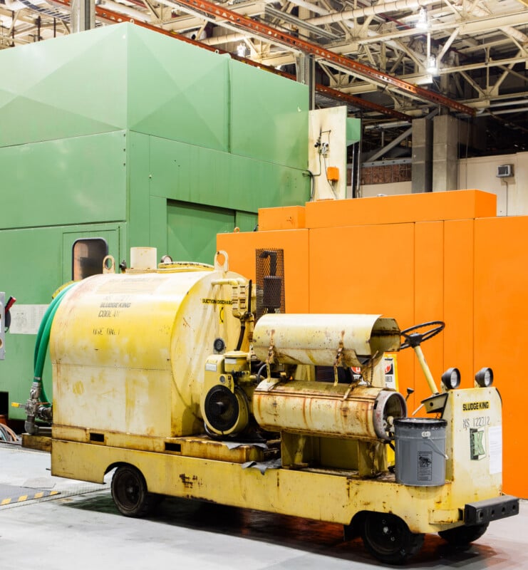 A yellow industrial floor cleaning machine stands in front of large green and orange factory equipment inside a brightly lit manufacturing facility.