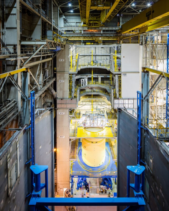 Interior of a large industrial facility with yellow metal platforms, walkways, and railings surrounding a cylindrical machine part being assembled by workers in a spacious, high-ceilinged area with multiple levels and equipment.