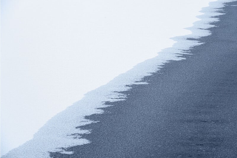 Abstract aerial view of a shoreline, with light, textured sand meeting darker water in an irregular, jagged line. The scene gives a minimalist, tranquil impression with soft, muted colors and gentle contrasts.