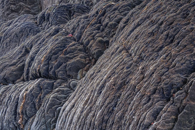 Close-up view of textured, dark gray volcanic rock with layered, wavy patterns and grooves, creating a rugged, rippled surface.