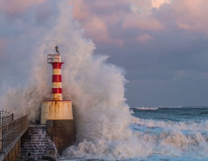 A large wave crashes against a red and white striped lighthouse on a concrete pier, sending spray high into the air. The ocean is rough, and the sky is filled with pink and gray clouds at sunset.