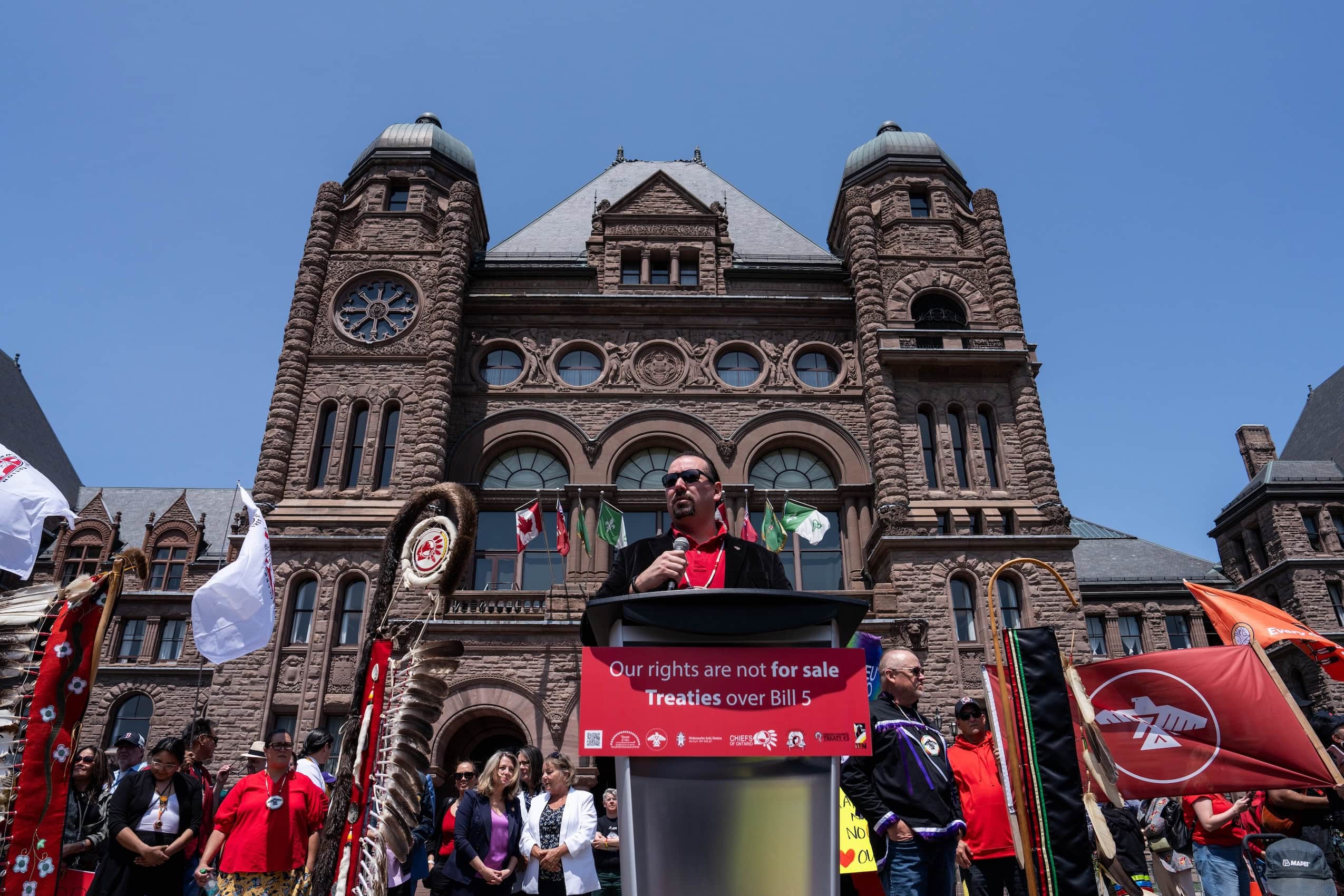 Ontario Regional Chief Abram Benedict stands at a lectern with a microphone and speaks to a crowd, with the provincial legislature in the background.