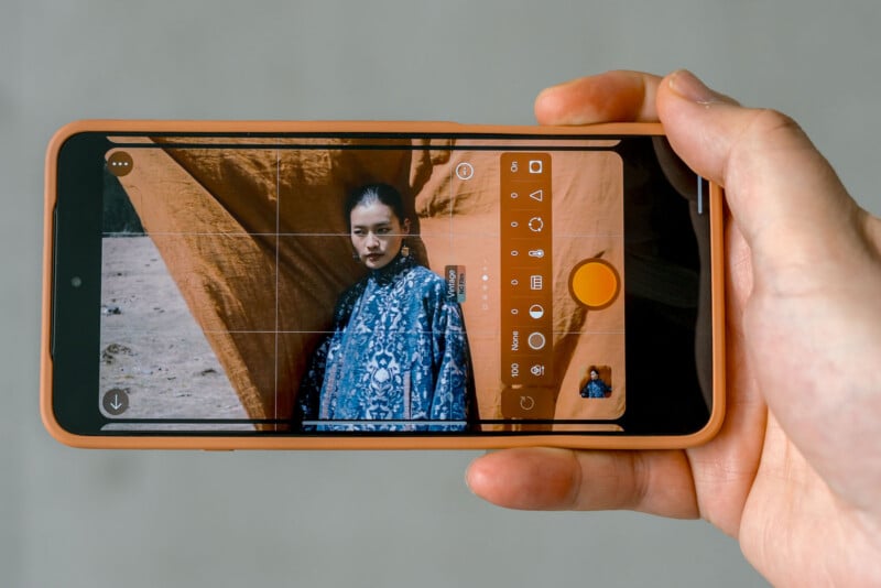 A hand holds a smartphone, taking a photo of a person in a blue patterned outfit standing in front of an orange fabric backdrop. The phone screen displays camera controls and the person being photographed.