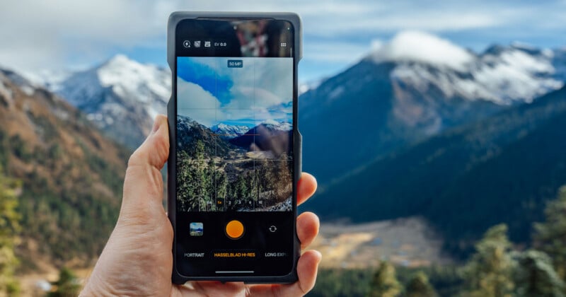 A hand holds a smartphone taking a photo of a scenic mountain landscape with snow-capped peaks, pine trees, and a partly cloudy sky in the background.