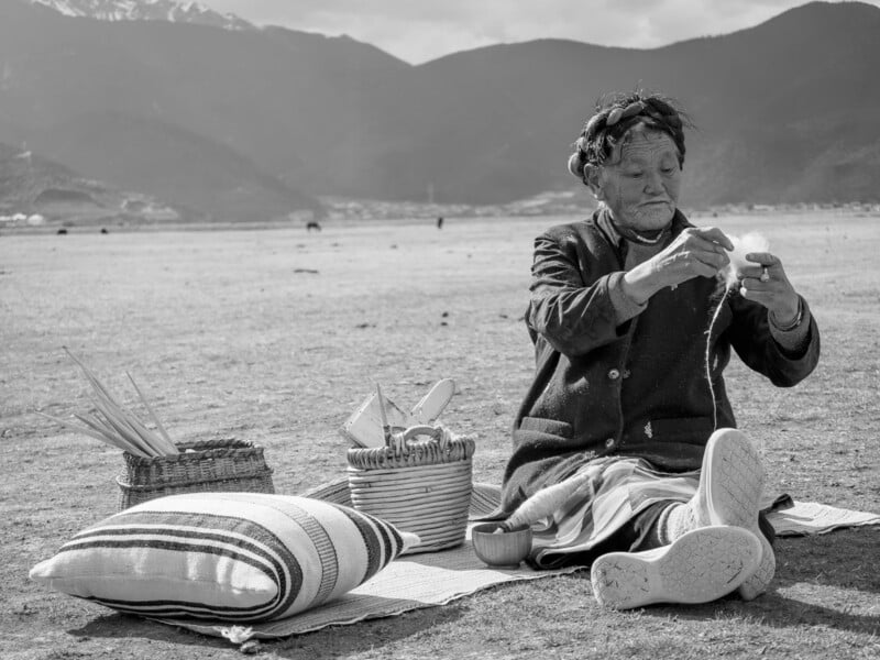 An elderly person sits on a mat outdoors with mountains in the background, weaving or handling string, beside baskets and a striped pillow. The setting appears rural and peaceful.