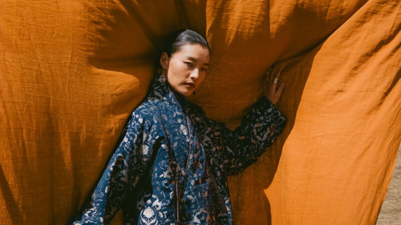 A person in a patterned blue robe stands against a backdrop of large, draped orange fabric, looking confidently at the camera with one hand resting on the fabric.