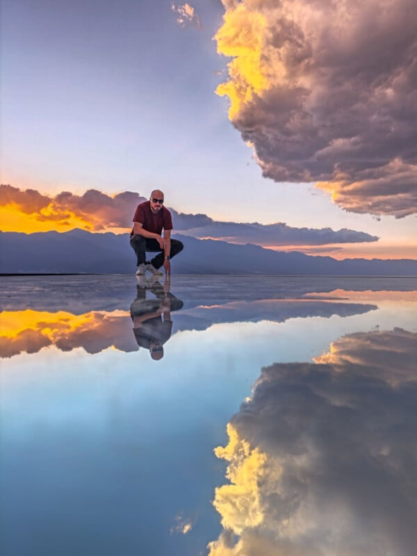 A person in sunglasses crouches on a reflective water surface, with clouds, mountains, and a vibrant sunset sky mirrored below them. The scene is colorful and serene.