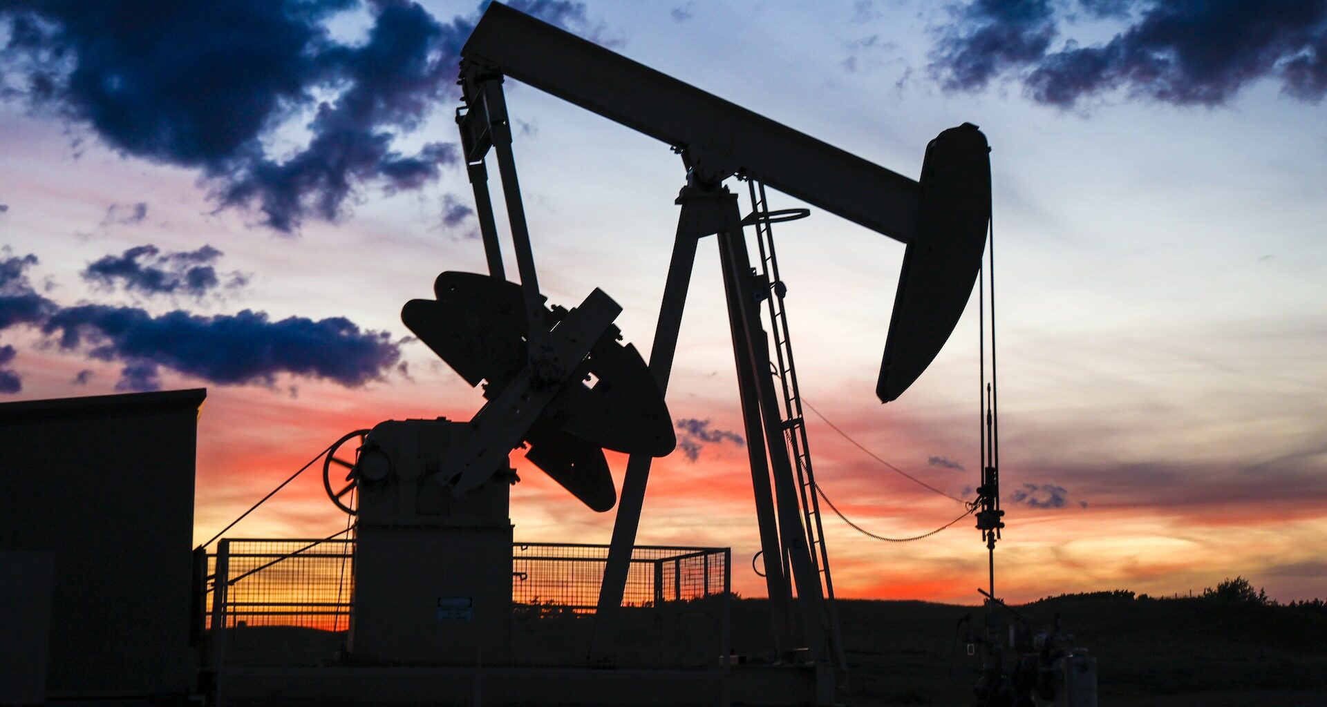 The image shows the silhouette of a pumpjack drawing out oil from a well head against an orange sunset.