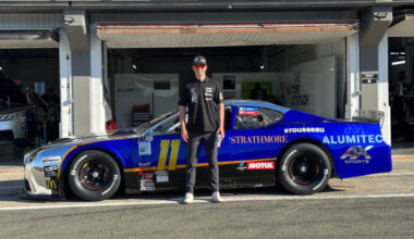 Person standing beside a blue race car in a pit garage, wearing a black shirt and cap, number 11 on the car.