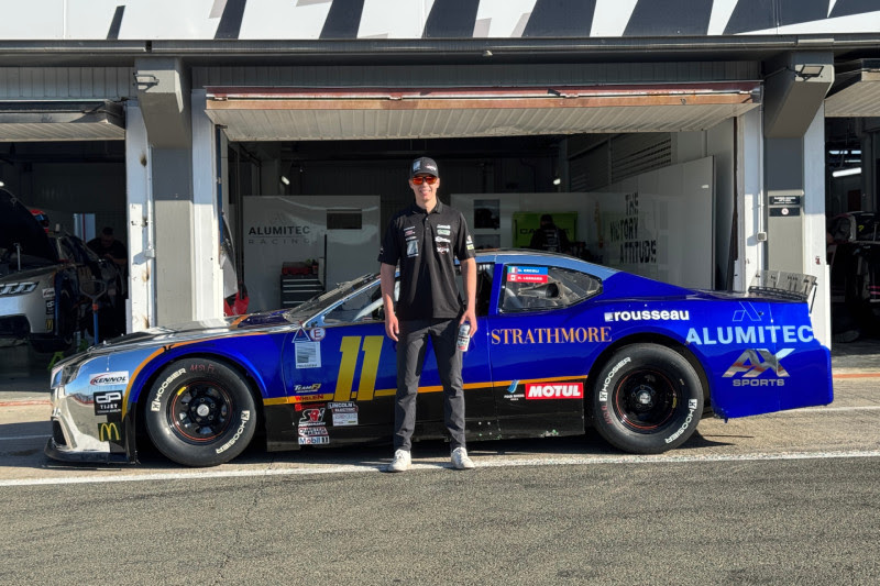 Person standing beside a blue race car in a pit garage, wearing a black shirt and cap, number 11 on the car.