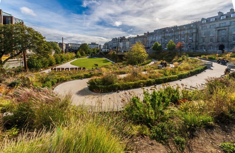 A landscaped urban park with curved pathways, grassy lawns, flower beds, and trees, surrounded by historic buildings under a partly cloudy sky. People are walking and sitting throughout the park.
