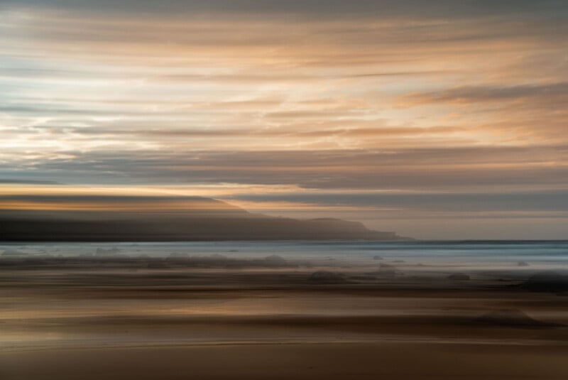 A blurred, dreamy view of a beach at sunset, featuring soft orange and blue hues in the sky, distant hills, and gentle waves washing over the sandy shore.