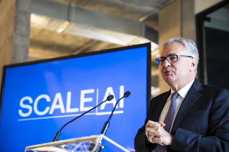 A side-angle shot of Julien Billot at a lectern. There is a large video monitor in the background displaying Scale AI's blue-and-white logo.