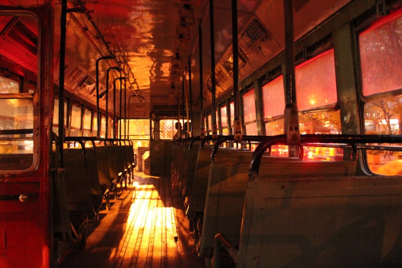 Interior of an empty, old bus at night, illuminated by warm, orange streetlights from outside the windows, creating dramatic shadows and reflections on the seats and floor.