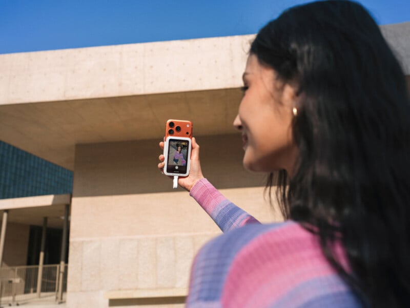 A woman with long dark hair smiles as she takes a selfie with her phone outside a modern beige building under a clear blue sky.